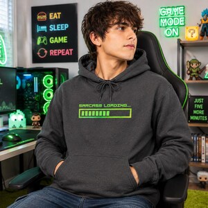 Könnte beinhalten: Ein dunkelgrauer Kapuzenpullover mit einer grünen, verpixelten Ladebalken-Grafik auf der Brust, über der der Text "SARCASM LOADING..." gedruckt ist. Der Hoodie verfügt über eine Kängurutasche auf der Vorderseite und passende dunkelgraue Kordeln.