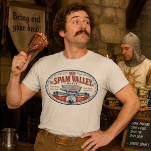 May include: A light gray t-shirt featuring a graphic oval logo that reads Spam Valley Meats, Breakfast, Lunch and Dinner, with an illustration of a pig, a crown, and a small figure in a helmet, all printed in red, blue, and white ink on the chest.