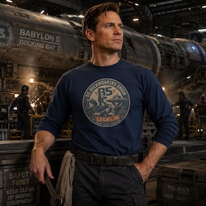 May include: A navy blue long-sleeve t-shirt featuring a circular graphic on the chest with the text B5 DOCKWORKERS UNION LOCAL 14, depicting a worker and a space station. The shirt is worn with dark trousers and a black belt in an industrial setting.