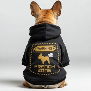 以下が含まれることがあります： 「WARNING FRENCH ZONE」と書かれた金色のグラフィックが入った黒い犬用パーカー。グラフィックはフレンチブルドッグのシルエットとガスの雲が特徴です。犬は茶色と白です。