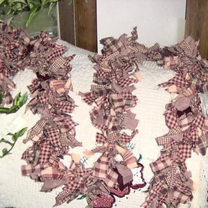 May include: A decorative garland made of fabric strips in various plaid patterns, primarily in shades of burgundy, cream, and tan. The garland is arranged in a wavy pattern, suggesting it could be used as a decorative accent for a home or event.