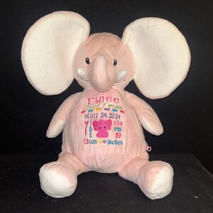 May include: A soft pink elephant toy with large white ears and embroidered details. The elephant features a personalized embroidered design on its belly, including a name, date, time, weight (3.18 kg), and length (48.26 cm).