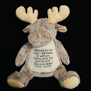 Puede incluir: Un alce de peluche marrón con grandes cuernos beige. El alce está sentado con las piernas cruzadas y tiene una mancha blanca en el pecho con el texto bordado: "Although we are apart. I will always be with you. Kisses. hugs, and love forever. Colten Jacob Zimdars 9/2023-11/2024"