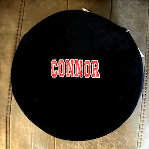 May include: Black round fabric patch with the name "CONNOR" embroidered in red letters with white outlines.