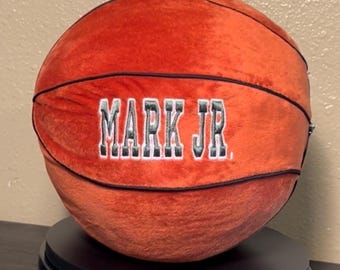 May include: A plush orange basketball with black stitching and the name "MARK JR." embroidered in white and gray lettering.