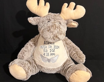 May include: A plush brown moose with tan ears and hooves. The moose has a white patch on its chest with the text "Asher March 1st, 2024 6lb 2oz 4:16 PM" embroidered in black. The patch also has a silver heart with wings embroidered on it.
