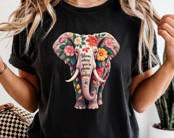 May include: Black t-shirt featuring a floral elephant design. The elephant is adorned with pink, yellow, and red flowers, with the text "Bloom where you're planted". The shirt has short sleeves and a crew neck.