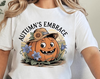 May include: White t-shirt featuring a smiling cartoon pumpkin wearing a straw hat with a sunflower. The pumpkin is surrounded by blue flowers and the text "Autumn's Embrace" in a circular design.