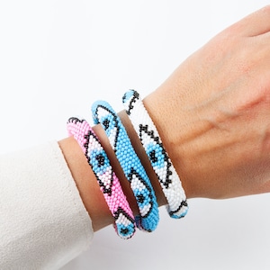May include: Three beaded bracelets with a white, light blue, and pink background. Each bracelet has a black and white eye design.