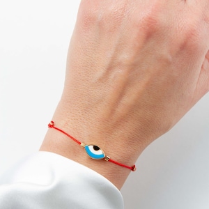 Puede incluir: Un brazalete de cuerda roja con un dije de ojo turco dorado. El ojo turco es azul, blanco y negro.