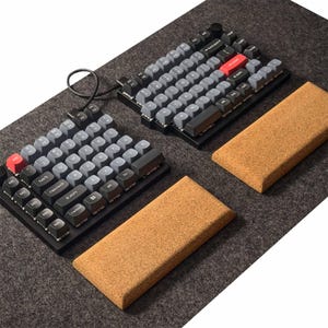 May include: Two black and gray mechanical keyboards with gray keycaps and red accent keys are displayed on a gray felt surface. Two rectangular, light brown cork wrist rests are also present. The keyboards are connected by a black cable.