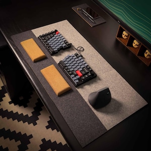 May include: A modern desk setup featuring two split ergonomic keyboards with gray and black keys, a red accent key, and two cork wrist rests. A gray ergonomic mouse and a smartphone are also visible.