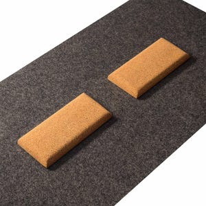 May include: Two rectangular cork blocks with rounded edges are positioned on a dark gray felt surface. The blocks have a natural, light brown color and a textured appearance. The felt surface provides a contrasting backdrop.