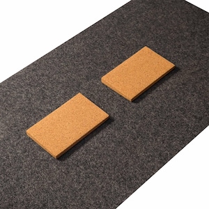 May include: Two rectangular cork coasters sit on a dark gray, textured surface. The coasters are a light brown color and appear to be made of natural cork material. The background is a dark gray, possibly felt, material.