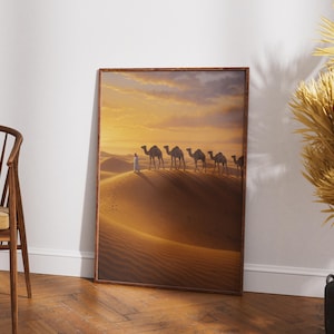 Puede incluir: Una impresión enmarcada representa una escena desértica con una fila de camellos y una figura vestida de blanco caminando sobre una duna de arena. El cielo es una cálida mezcla de tonos naranja y amarillo, sugiriendo una puesta de sol. El marco es marrón.