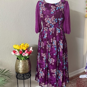 May include: A purple floral midi dress with a round neckline and 3/4 sleeves. The dress has a gathered skirt and a fitted bodice.