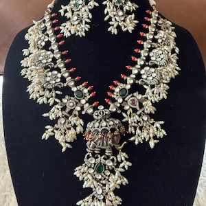 May include: An ornate necklace and matching earrings set. The jewelry features a silver-toned metal with intricate detailing, including small white pearls, red and green accents, and elephant motifs. The necklace has a central pendant with a large green stone.