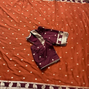 May include: A burnt orange saree featuring a repeating gold dot pattern and a wide dark maroon border with gold elephant and peacock motifs. A matching dark maroon blouse with gold trim and similar gold elephant and peacock patterns is folded on top.