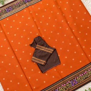 May include: An orange saree featuring a repeating pattern of small white floral motifs. The fabric has a wide dark brown border decorated with a colorful floral and peacock design. A matching dark brown blouse piece with striped trim is folded in the center.