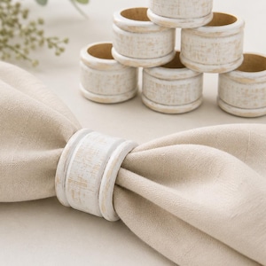 May include: A set of six cylindrical napkin rings with a distressed white painted finish over a natural wood base. One ring is shown holding a folded beige linen napkin, while the other five are stacked in a pyramid formation on a flat surface.