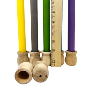 May include: A collection of colorful taper candles in yellow, gray, green, and purple, each set in a small, unfinished wooden holder. A wooden ruler is placed next to the candles. The candles are approximately 8 inches tall.
