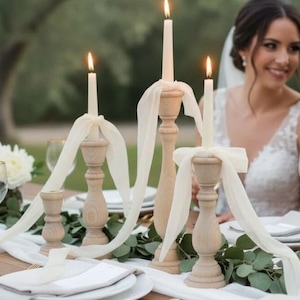 Puede incluir: Una mesa de boda con tres candelabros de madera, cada uno con una vela encendida y cubierto con una cinta blanca. La mesa está puesta con platos blancos, vegetación y una pareja en el fondo.