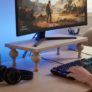 Op de afbeelding: Een lichtgekleurde houten monitorstandaard met vier decoratieve, gedraaide poten. De standaard is rechthoekig en ondersteunt een zwarte computermonitor met een videogame. Een zwart toetsenbord en een koptelefoon zijn ook zichtbaar.