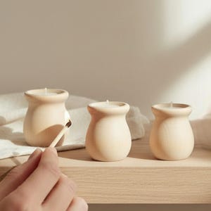 May include: Three beige, jar-shaped candles sit on a wooden shelf. A hand holds a lit match near one candle. The candles have a smooth, rounded design and a small wick at the top. The background is a neutral color.