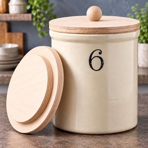 May include: A cylindrical cream-colored ceramic canister featuring the black number 6 printed on the front. The canister includes a light-colored wooden lid with a round knob handle and a separate matching wooden lid resting beside it.