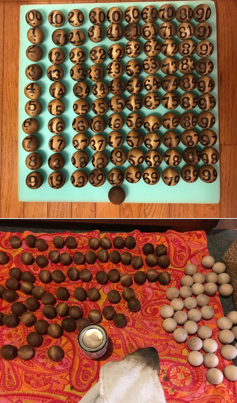 May include: A set of 90 wooden balls with black numbers painted on them. The balls are arranged in a grid on a light blue surface. The balls are used for a game of bingo.