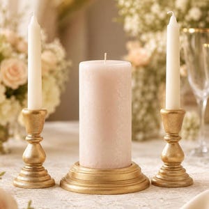 May include: Two slender white taper candles in gold-toned metallic candle holders flank a single, thick, cylindrical pale pink pillar candle resting on a matching circular gold-toned base. The set features a consistent matte gold finish.