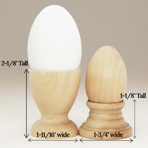 Puede incluir: Dos hueveras de madera. Una sostiene un huevo blanco y mide 5,4 cm de alto y 4,3 cm de ancho. La otra es un huevo de madera, de 2,8 cm de alto y 4,4 cm de ancho. Ambas son de madera clara.