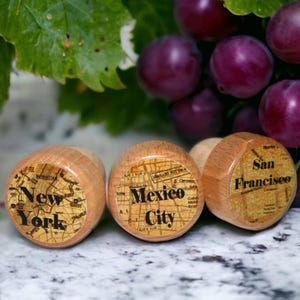 May include: Three cylindrical wooden bottle stoppers featuring circular inlays with vintage-style map prints. Each inlay displays a city name in black text: New York, Mexico City, and San Francisco, set against a detailed grid of street lines.