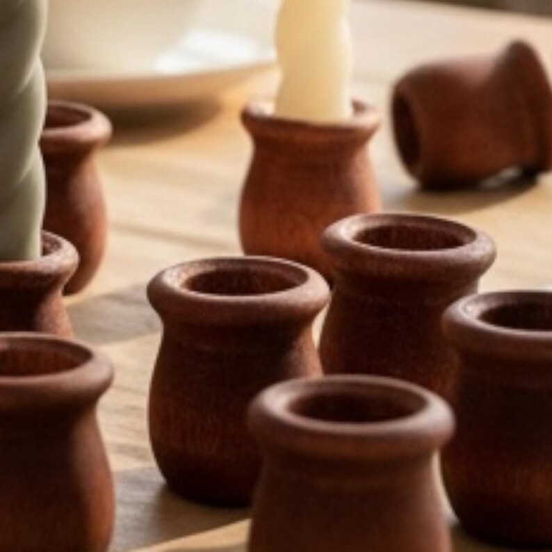 May include: A collection of small, terracotta-colored candle holders. The holders have a rounded shape with a slightly flared rim. Some hold white or green candles. The holders are arranged on a light-colored wooden surface.