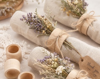May include: Light beige fabric napkins rolled and secured with smooth wooden napkin rings. Each bundle is decorated with a small bouquet of dried lavender and white flowers tied with twine. One napkin features a brown paper tag with the text thank you.