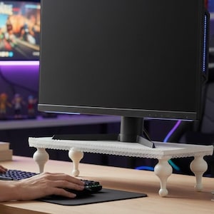 Op de afbeelding: Een witte monitorstandaard met vier decoratieve, afgeronde poten. De standaard heeft een kanten rand en ondersteunt een zwarte computermonitor. De hand van een persoon is zichtbaar die een toetsenbord en muis op een bureau gebruikt.