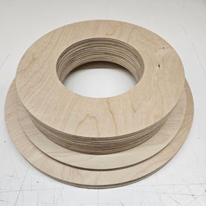 May include: A stack of unfinished wooden circles with a hole in the center. The wood has a light, natural color and visible grain. The circles are of varying sizes, with a few larger ones at the bottom.
