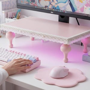 Op de afbeelding: Een roze monitorstandaard met een witte kanten rand en parelaccenten. De standaard heeft vier decoratieve, afgeronde poten. Een wit toetsenbord met roze en blauwe toetsen, een roze muis en een wolk-vormige muismat liggen op het bureau.