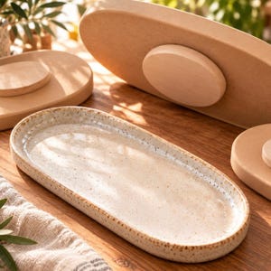 May include: An oval ceramic tray with a speckled cream glaze and a raised rim. The tray is surrounded by several light-colored wooden circular and oval bases with smooth, matte surfaces, arranged on a wooden surface with natural light.