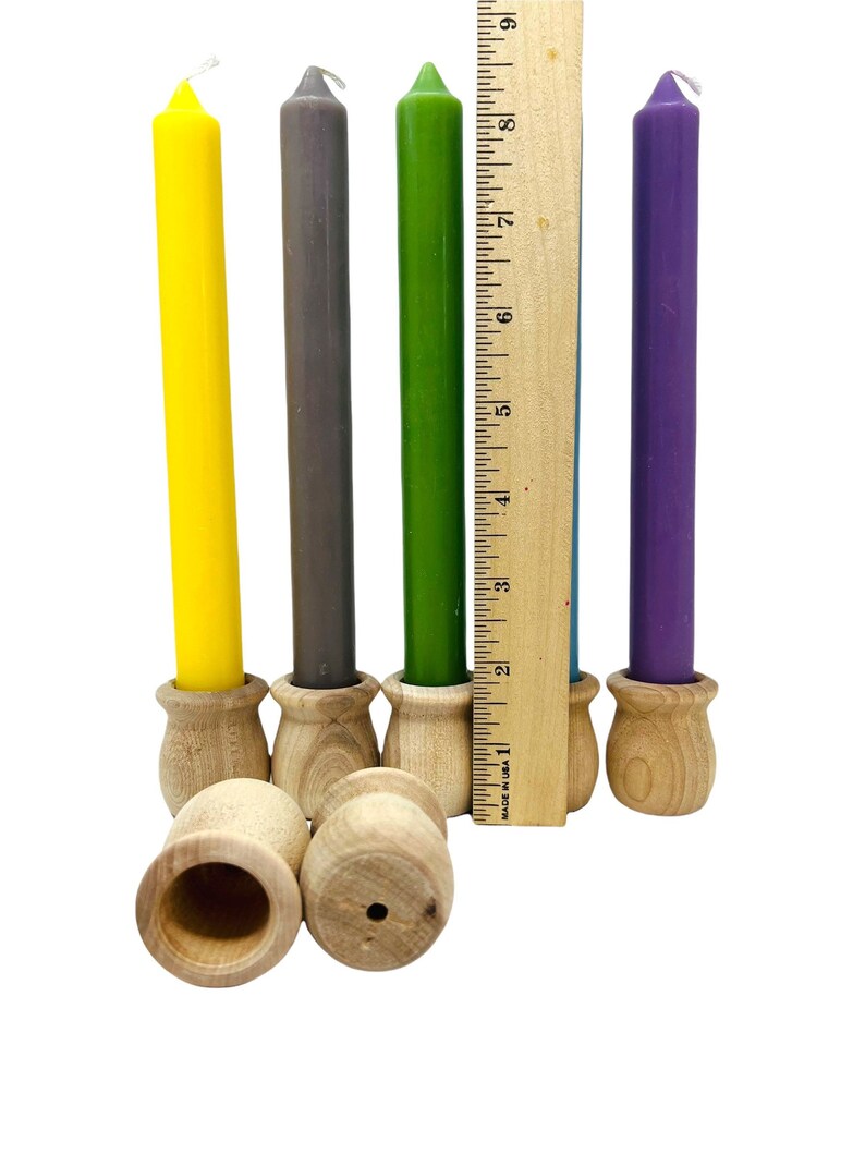 May include: A collection of colorful taper candles in yellow, gray, green, and purple, each set in a small, unfinished wooden holder. A wooden ruler is placed next to the candles. The candles are approximately 8 inches tall.
