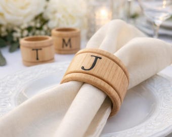 May include: A light-coloured wooden napkin ring featuring a dark engraved letter J, holding a folded cream-coloured fabric napkin on a white plate. Two additional matching wooden napkin rings with the letters T and M are visible in the background.