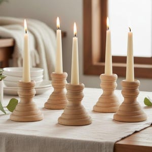 May include: Five light-colored wooden candle holders with a tiered, rounded base design. Each holder supports a single, slender, off-white taper candle. The candles are lit, emitting a small flame at the top of each wick, arranged on a table.