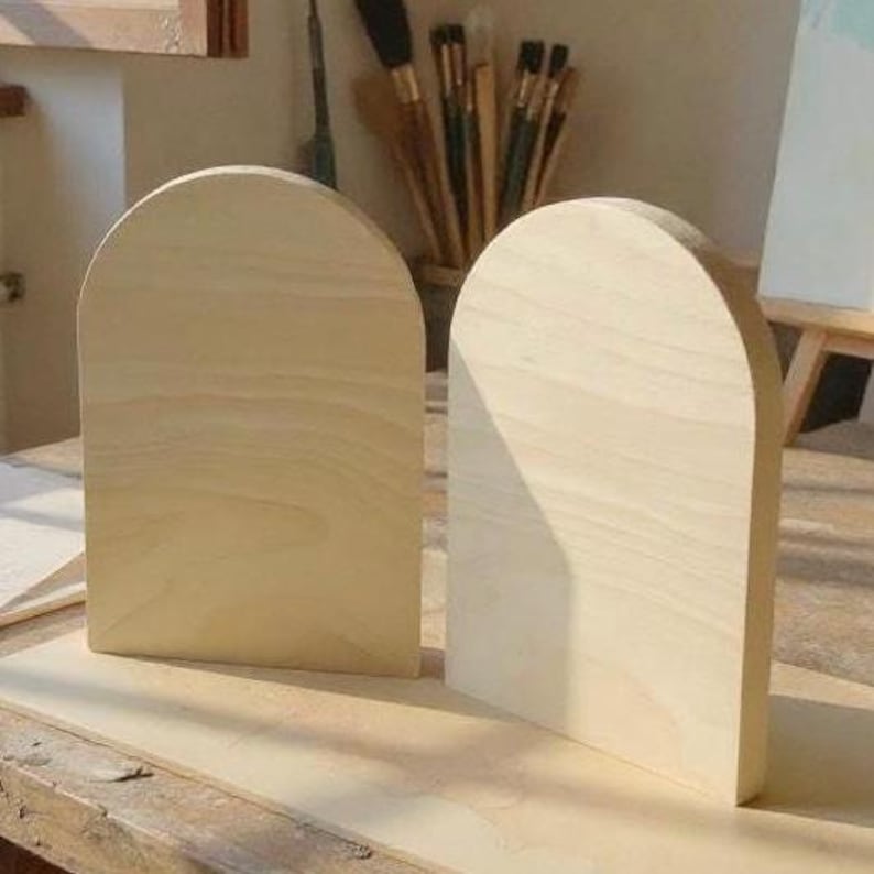 Puede incluir: Dos placas de madera clara sin terminar, cada una con una superficie lisa y plana y una parte superior en forma de arco redondeado. Estas dos piezas de madera id&eacute;nticas se mantienen en posici&oacute;n vertical, mostrando una veta natural en tonos beige.