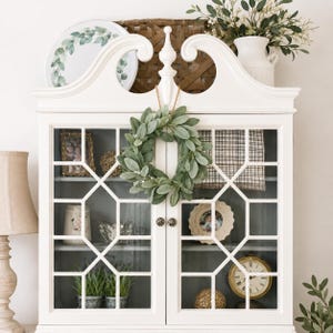 May include: A white wooden cabinet with glass doors featuring a geometric grid pattern. A small green leaf wreath hangs from the center top. Inside, the shelves display various decorative items including a clock, picture frames, and plants.