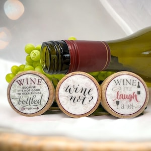May include: Three round wooden wine bottle stoppers with printed designs. One reads 'WINE BECAUSE IT'S NOT GOOD TO KEEP THINGS bottled UP', another 'wine not?', and the third 'WINE A LITTLE laugh a lot' with small wine glass and bottle icons.