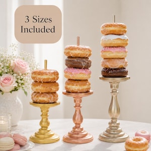 May include: Three metallic pedestal stands of varying heights, each featuring a central vertical rod for stacking donuts. The stands are finished in gold, copper, and silver tones. Text in the upper left corner reads 3 Sizes Included.