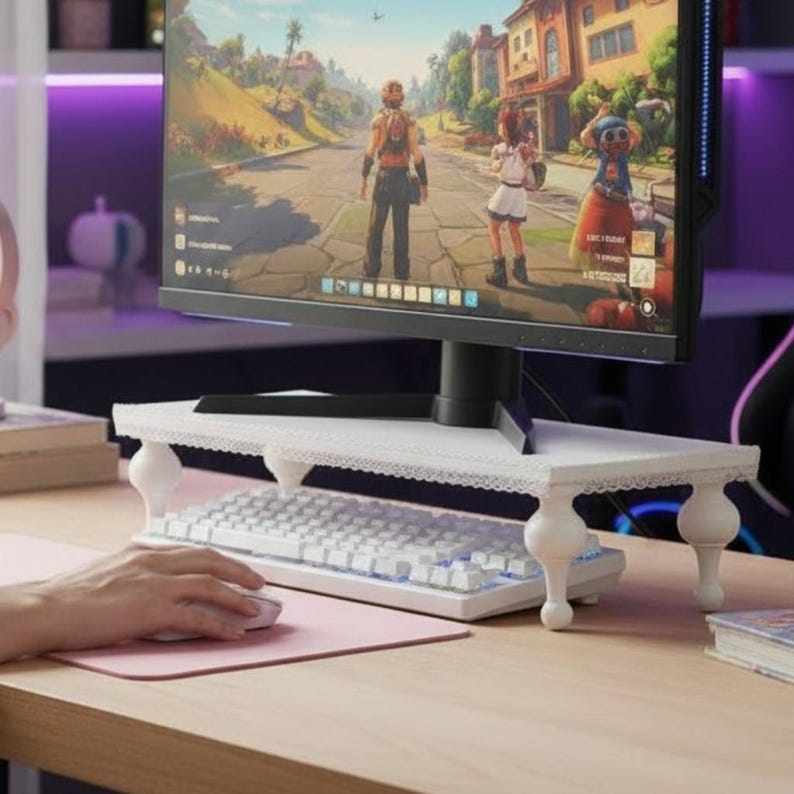 Op de afbeelding: Een witte monitorstandaard met een kanten rand en vier afgeronde poten. Onder de standaard staat een wit toetsenbord en een computermonitor toont een videogame. Een hand gebruikt een witte muis op een roze muismat.