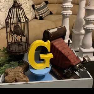 May include: A yellow wooden letter G sits on a blue pedestal. A vintage camera sits on a wooden tray with a birdcage and a butterfly.