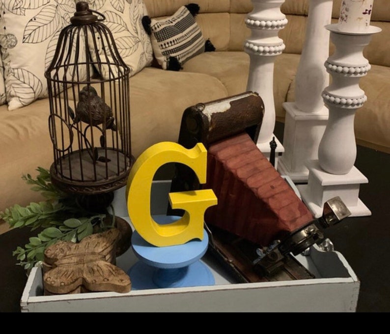 May include: A yellow wooden letter G sits on a blue pedestal. A vintage camera sits on a wooden tray with a birdcage and a butterfly.