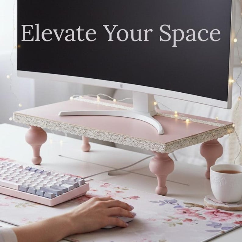 Op de afbeelding: Een roze monitorstandaard met een kanten rand en vier decoratieve poten. De standaard ondersteunt een witte monitor en heeft de tekst "Elevate Your Space" op het scherm. Een toetsenbord en muis staan op de voorgrond.